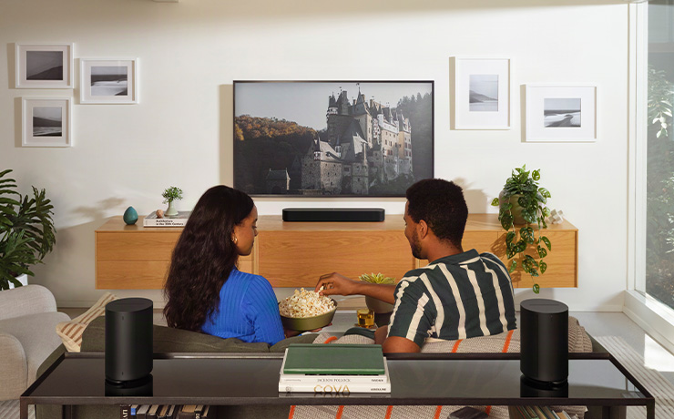 A pair of black Sonos Era 100 SL speakers on a unit that sits behind a sofa