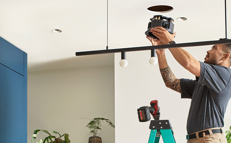 A man about to place the speaker into the cut-out hole in the ceiling.  He's on a green stepladder with a drill at the top of it.  He's wearing beige trousers and a dark grey top