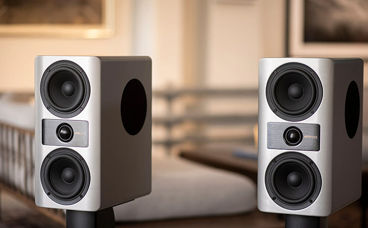 A pair of light grey speakers on stands with a blurred background of a living space