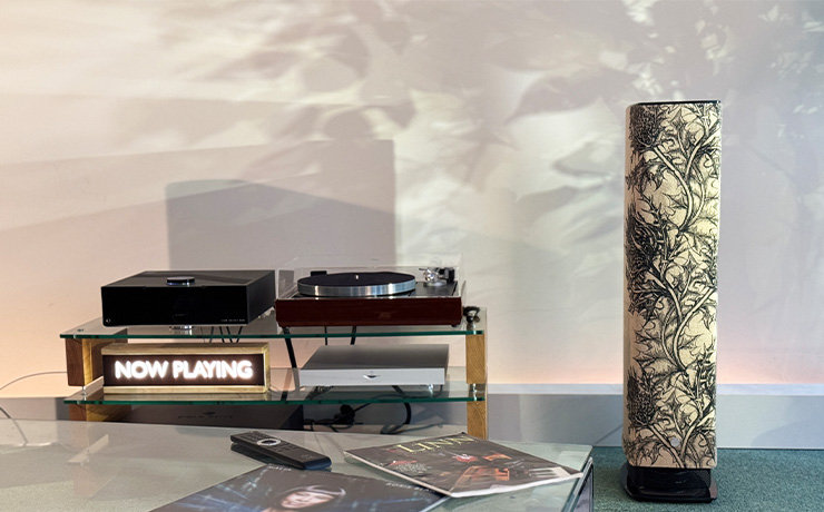 One of the ripcaster 530 Thistle speakers in the ripcaster showroom. there's an album and remote control in the foreground on a table. Beside the speaker is an LP12 and a DSM. There are many layers of shadow on the back wall.