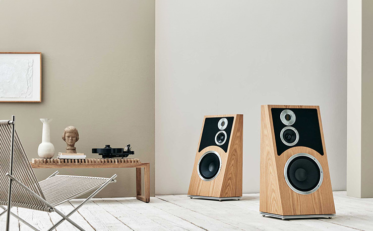A pair of Audiovector Trapeze speakers in Oak on the right of the image. On the left is a chair and at the back of the image a low table with a vase, a ceramic head and a turntable on