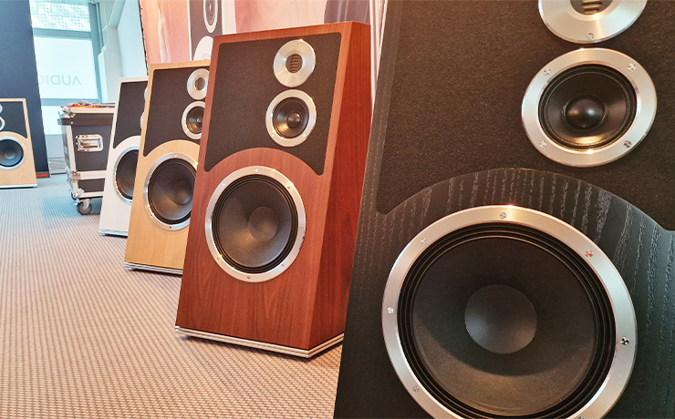 Five Audiovector Trapeze speakers lined up at the HiFi Show in Munich. Showing the four different finishes. Black at the front, walnut to the left and back, oak to the left and back and white to the left and back. Then one Oak one at the back. the carpet is a light beige with darker beige stripes