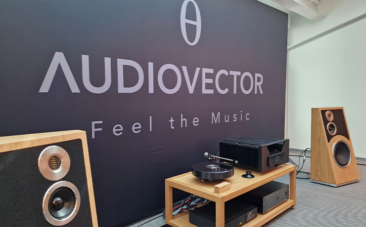 A pair of Audiovector Trapeze speakers in oak either side of some other HiFi equipment on a low unit. there's a large Audiovector board behind. This photo was taken at the Munich HiFi Show in 2024