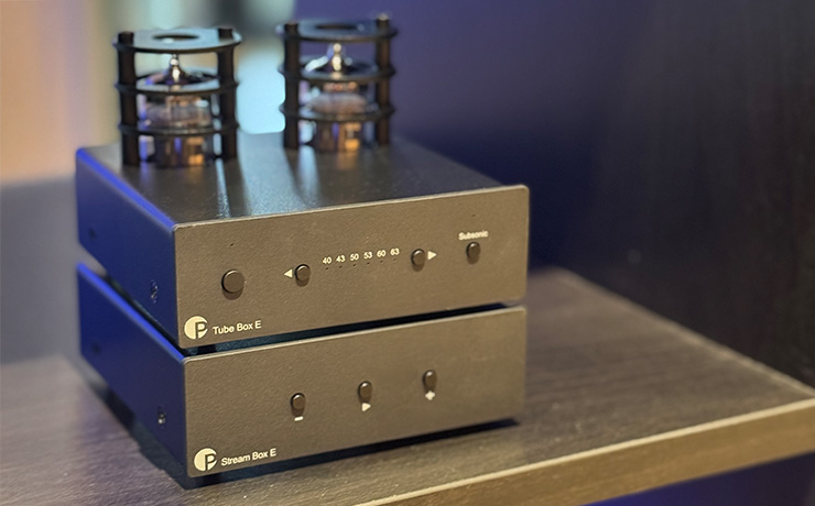 The Project Tube Box E on top of the Project Stream Box E on a wooden shelf with a blurred view out of a window