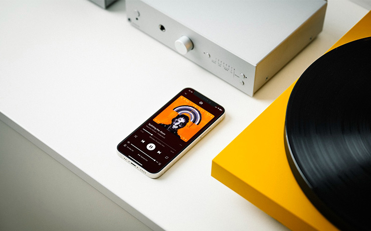 The Project Uni Box S3 amplifier in silver on a white surface with a phone in front of it and a yellow turntable beside it.