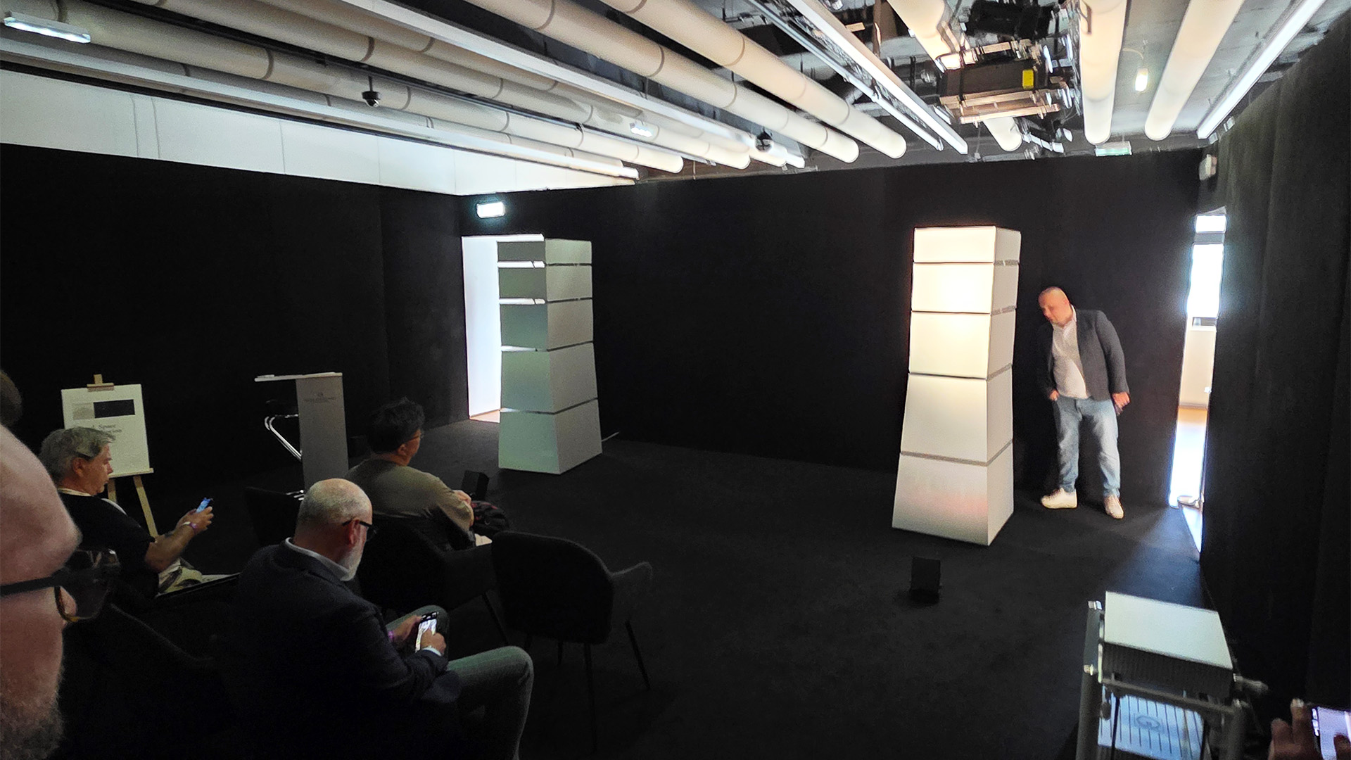 A room with a pair of white floorstanding speakers with people sitting viewed from the back