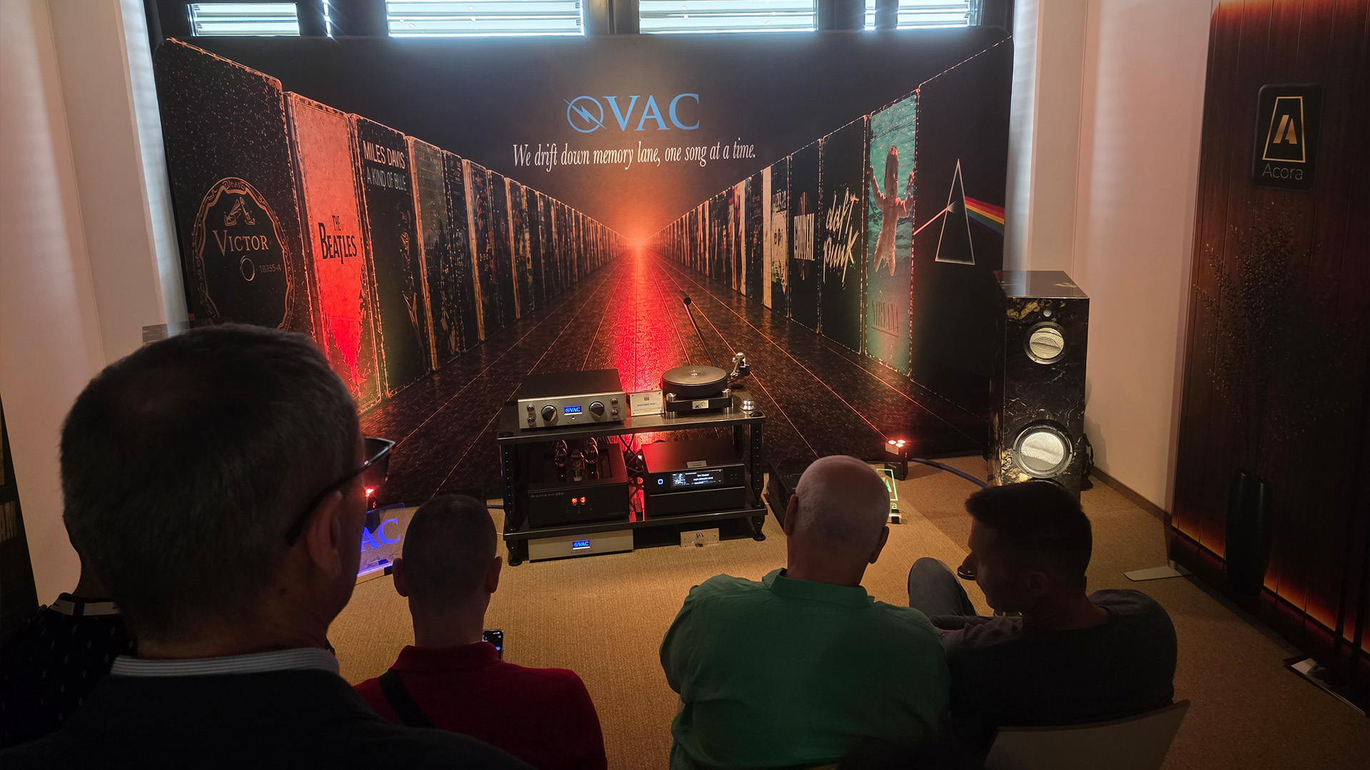 In the foreground are people facing away from us listening to the HiFi equipment at the front of the room