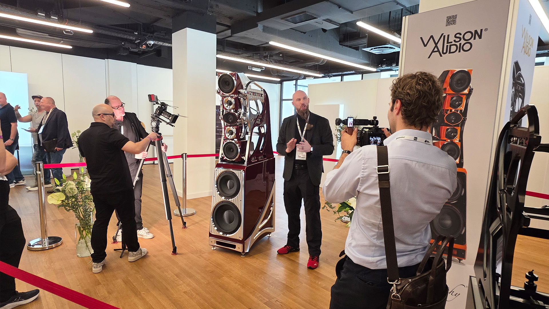 The Wilson Audio man beside a large floorstanding Wilson speaker.  There are two people filming him and other people standing around chatting
