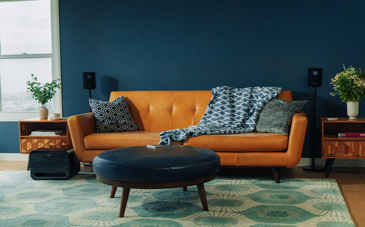 A pair of WiiM Sound speakers on stands either side of a leather sofa with a leather table or footstall in the centre