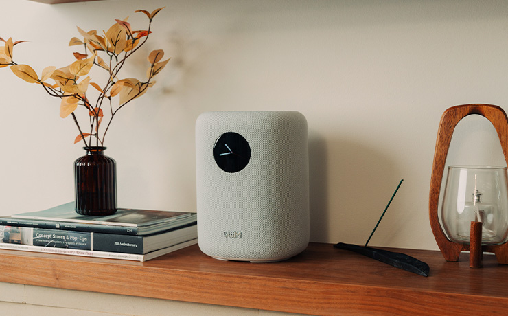 The WiiM Sound speaker on a wooden shelf with a light one side and a stack of books the other side with a vase on top of the books