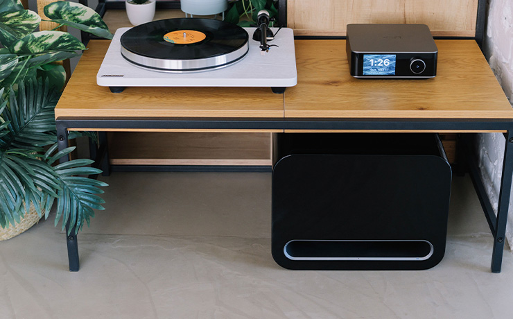 The WiiM sub pro in black underneath a desk that has a record player and a WiiM Ultra Streamer. There's an indoor plant to the left of the image, beside the desk