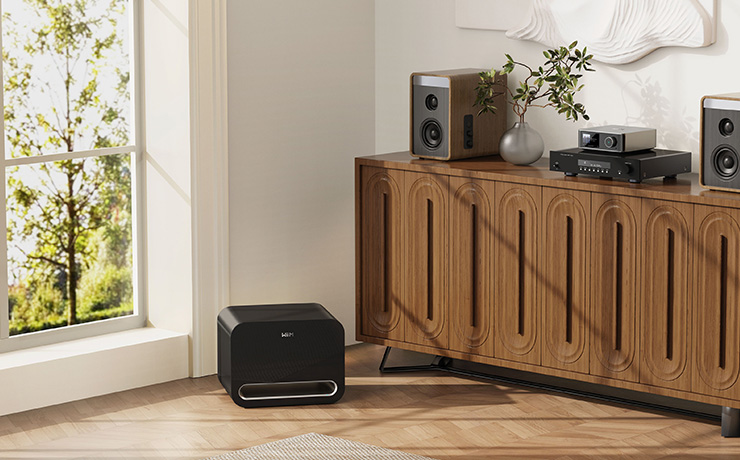 The WiiM Sub Pro in black on a wooden floor beside a large window. To the right is a wooden sideboard with various pieces of audio equipment on top