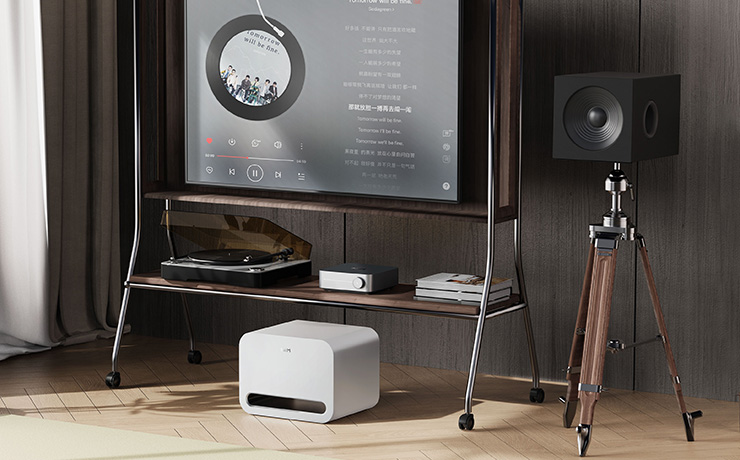 The WiiM Sub Pro in white on the floor of a living space with a large tv screen behind it and various pieces of audio equipment on a shelf. There's also a speaker on a tripod style stand to the right of the image.