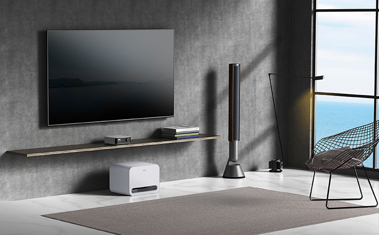 The WiiM Pro Sub in white on a floor in a living space underneath a shelf that has a small WiiM unit. There's a tv on the wall and a floorstanding speaker with a large window looking out to sea on the right along with a wire chair