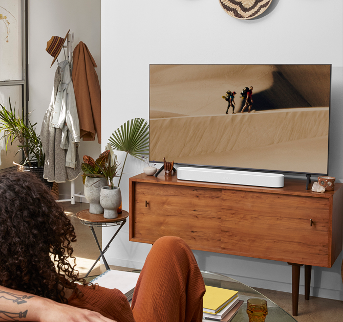 SONOS Beam (Gen 2) in white on a wooden tv cabinet.  there's a woman in the foreground with a man's arm along the sofa behind her.  the tv cabinet has a small table beside it with two vases containing foilage.  There's a hat stand to the side of the tv.  the tv is showing two people walking on sand dunes.