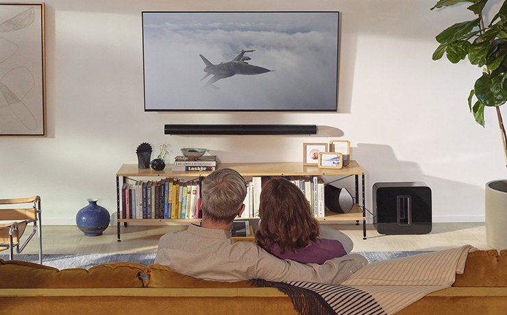 Sonos Sub on the floor in a living space beside a low unit. In the foreground is a couple sitting on a sofa viewed from behind
